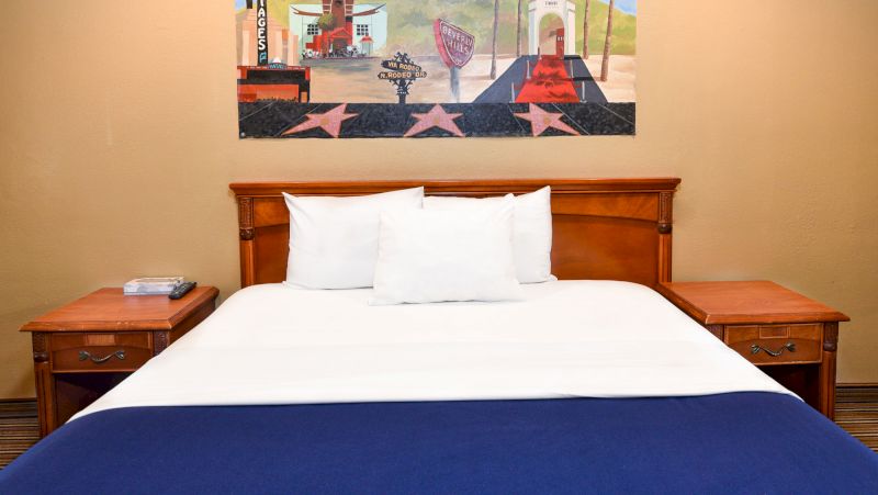 A hotel room bed with a wooden frame, white pillows, and a blue blanket at the foot, with a painting of the Hollywood Walk of Fame on the wall.