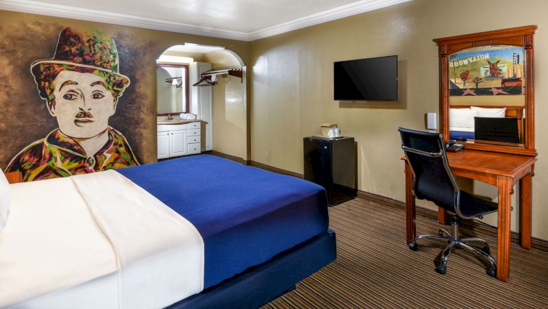 A hotel room with a blue bed, wooden desk, and a large mural of Charlie Chaplin on the wall.